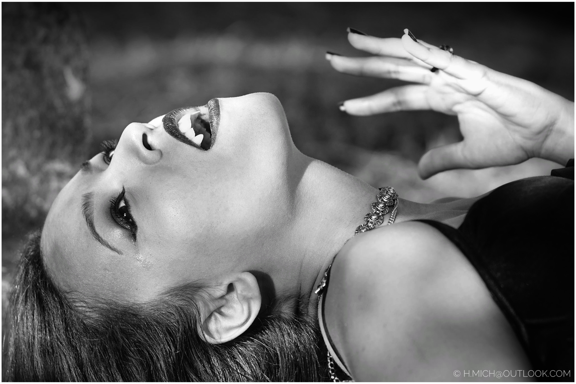 Diarium.Studio-One.Art.Br A black and white image of a person lying down with their head tilted back. They have dark nail polish, lipstick, and a choker necklace. The lighting highlights their facial features and the texture of their hair. (c) H.Mich