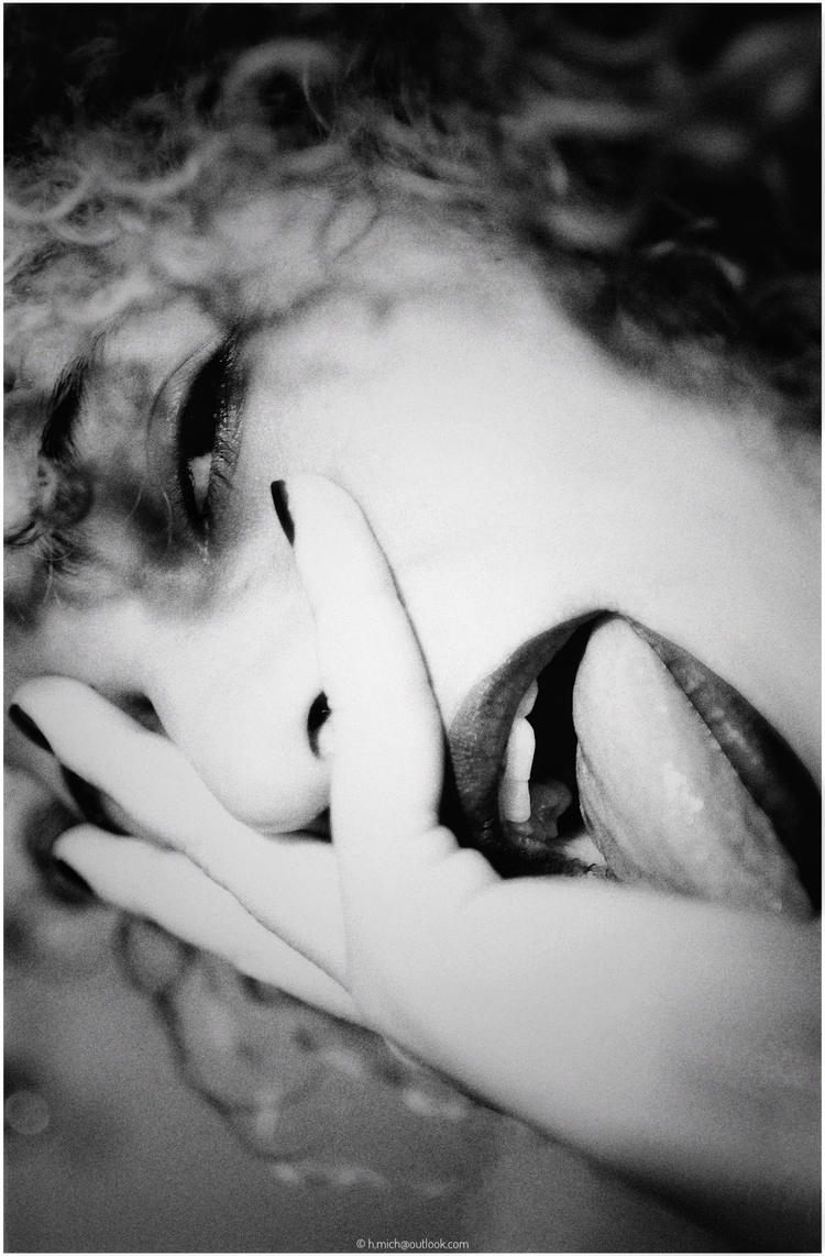 Diarium.Studio-One.Art.Br A close-up, black and white image of a person with curly hair. Their face is partially obscured by their hand, and they have their tongue out, touching their palm. Their expression is playful and whimsical. (c) H.Mich