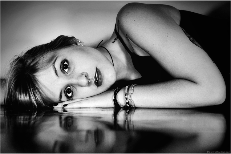 Diarium.Studio-One.Art.Br Black and white photo of a woman lying on her side, resting her head on the floor. She looks directly at the camera with a thoughtful expression. Her hand is adorned with bracelets, and she wears a nose ring. (c) H.Mich