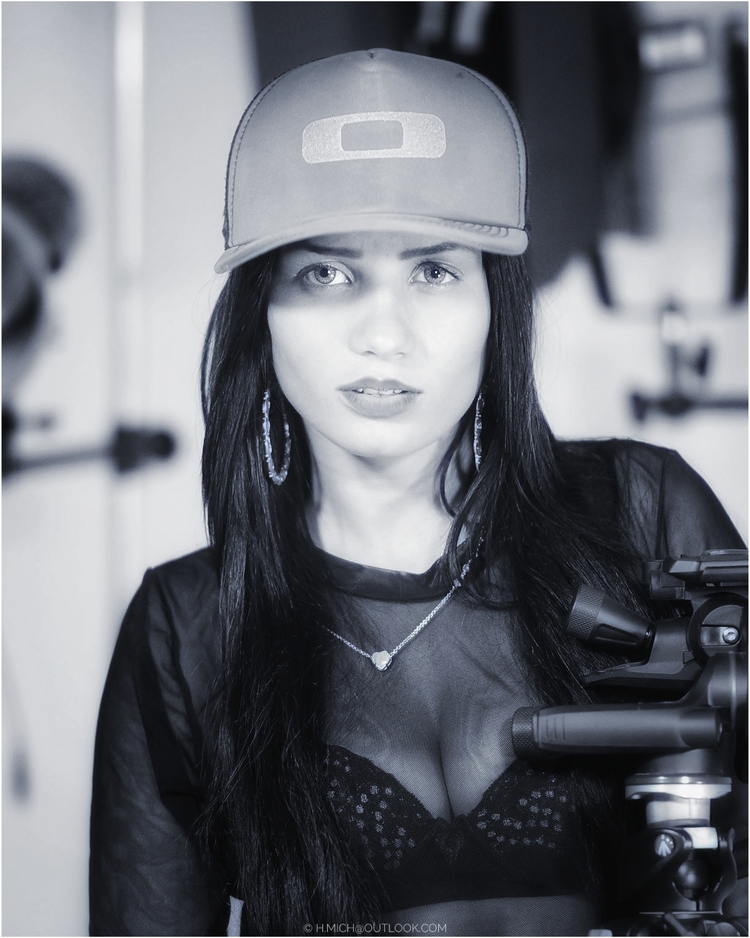 Diarium.Studio-One.Art.Br A woman with long dark hair wears a sheer black top and a cap. She has hoop earrings and a necklace with a heart pendant. The background is blurred, and theres a camera tripod partially visible. The image is in black and white. (c) H.Mich