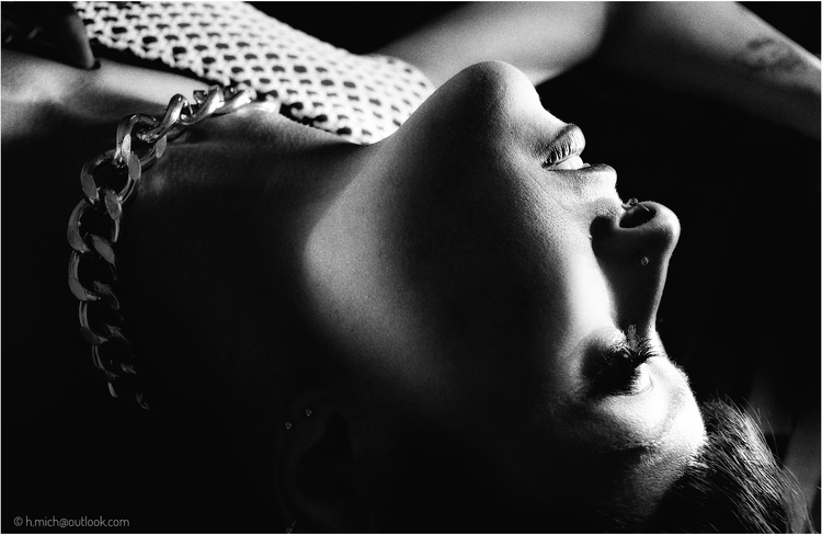 Diarium.Studio-One.Art.Br Black and white photo of a person lying on their back, head tilted upwards. They wear a chunky chain necklace and have a nose piercing. Light and shadows highlight their facial features, creating a dramatic effect. (c) H.Mich