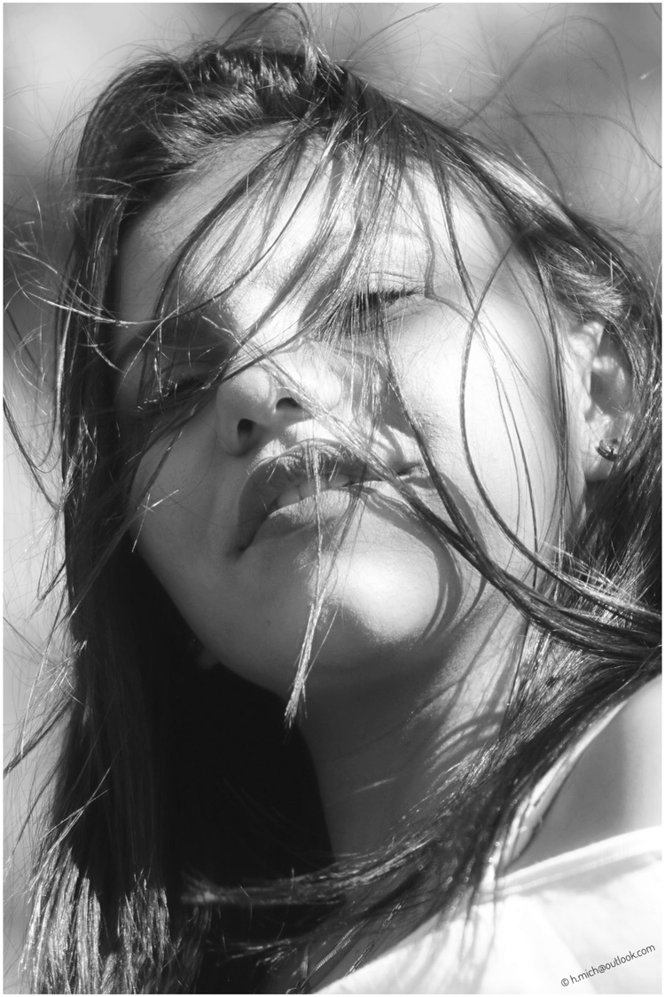 Diarium.Studio-One.Art.Br Black and white close-up of a woman with windblown hair partially covering her face, softly lit with shadows. She gazes upward, creating a serene and contemplative mood. (c) H.Mich