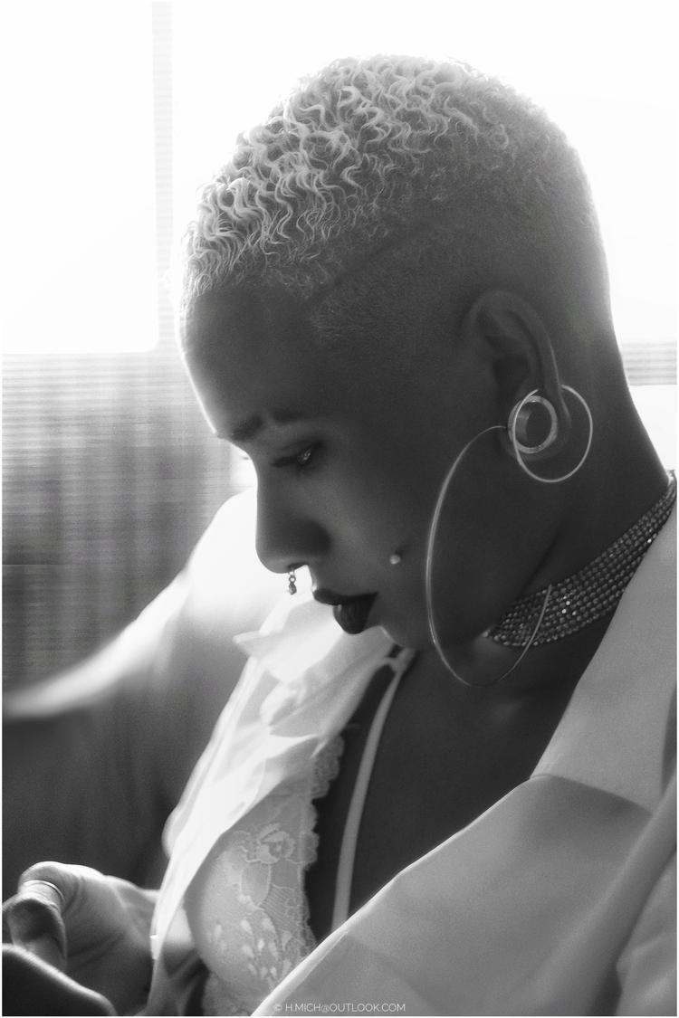 Diarium.Studio-One.Art.Br Black and white image of a person with short, curly hair, large hoop earrings, and a choker looking downward. They are wearing a blazer and lace top, seated in a softly lit environment. (c) H.Mich