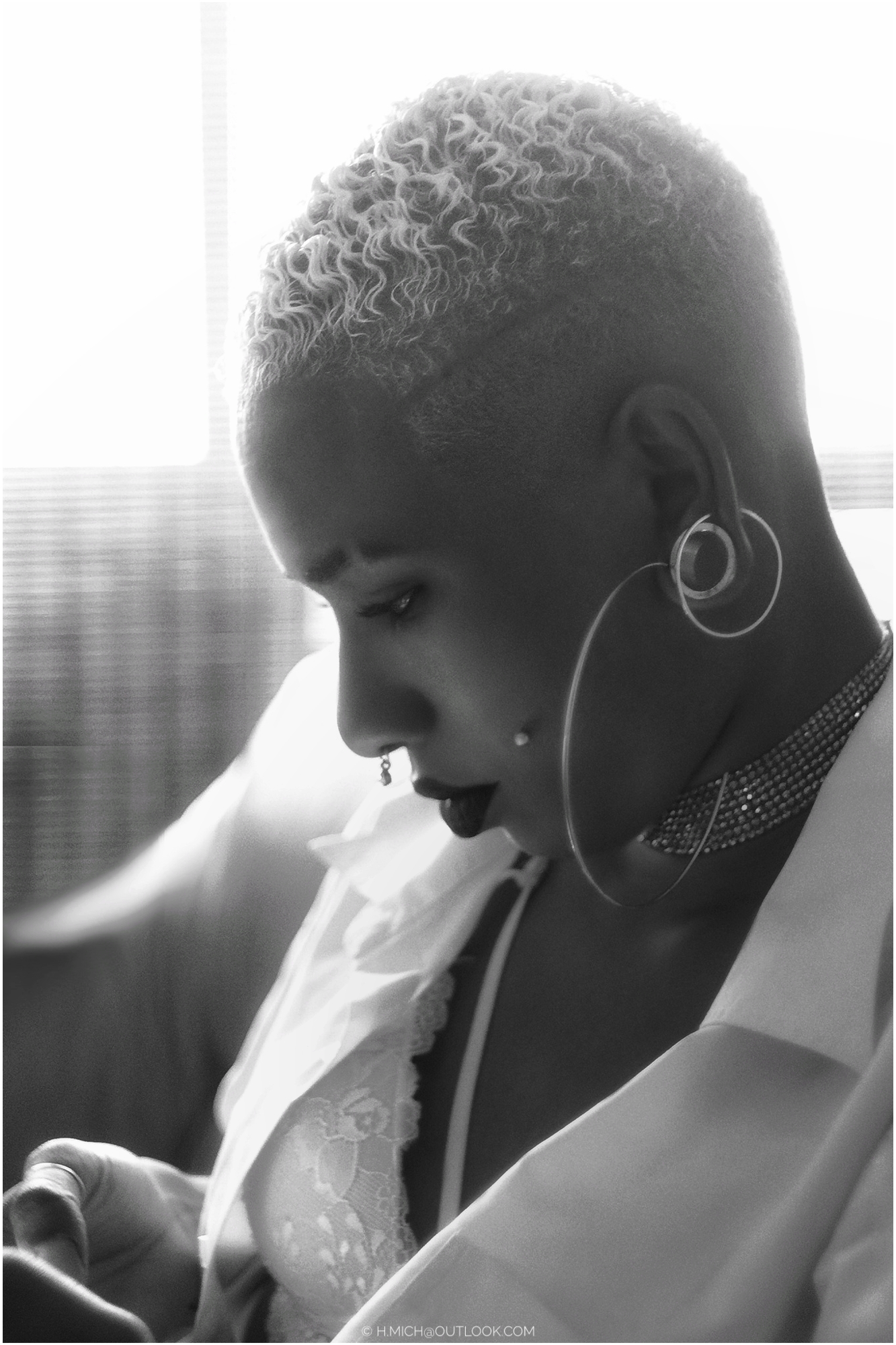 Diarium.Studio-One.Art.Br Black and white image of a person with short, curly hair, large hoop earrings, and a choker looking downward. They are wearing a blazer and lace top, seated in a softly lit environment. (c) H.Mich