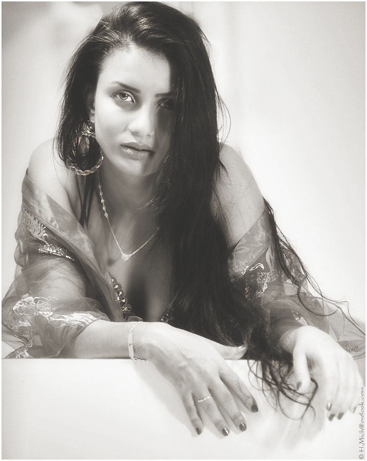 Diarium.Studio-One.Art.Br A woman with long dark hair leans forward, resting her arms on a surface. She wears hoop earrings, a necklace, and a sheer shawl over her shoulders. The image is in black and white, giving it a classic, timeless feel. (c) H.Mich