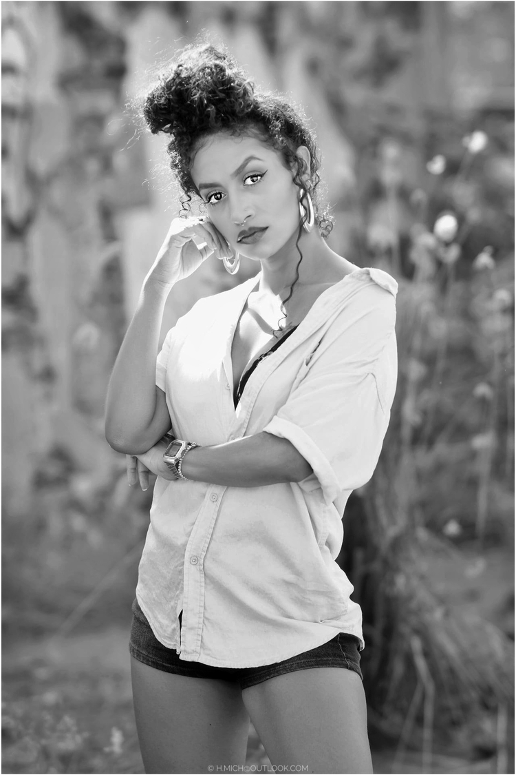 Diarium.Studio-One.Art.Br A woman with curly hair in a bun is standing in an outdoor setting. Shes wearing a loose shirt and shorts, looking directly at the camera with her hand resting on her cheek. The image is in black and white. (c) H.Mich