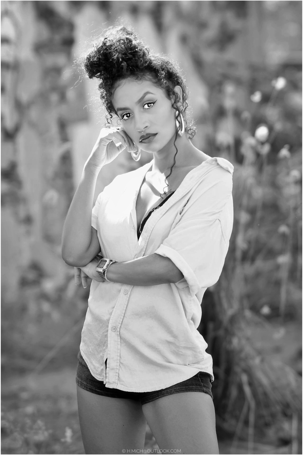 Diarium.Studio-One.Art.Br A woman with curly hair in a bun is standing in an outdoor setting. Shes wearing a loose shirt and shorts, looking directly at the camera with her hand resting on her cheek. The image is in black and white. (c) H.Mich