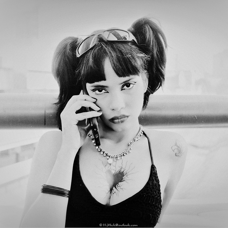 Diarium.Studio-One.Art.Br A person with dark pigtails holds a phone to their ear, wearing sunglasses on their head, a necklace, and a black top. The image is in black and white and includes a visible tattoo on their arm. (c) H.Mich