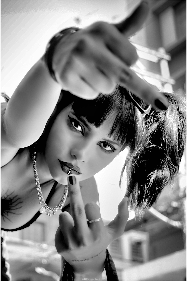 Diarium.Studio-One.Art.Br A black-and-white photo of a person with dark lipstick and nail polish, hair styled with bangs and pigtails, posing with a confident expression. They are giving a gesture with both hands, wearing a necklace, outdoors. (c) H.Mich