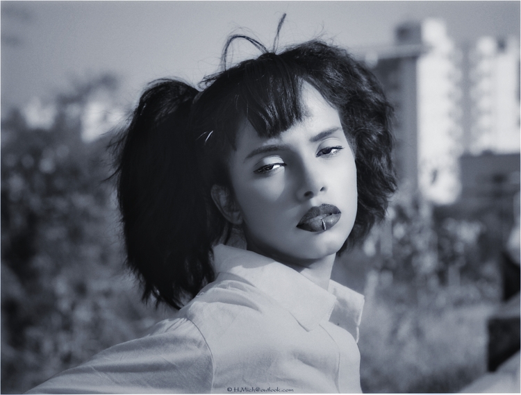 Diarium.Studio-One.Art.Br Black and white portrait of a person with dark hair in pigtails, wearing a light-colored shirt. They have a serious expression, with blurred buildings and trees in the background. (c) H.Mich