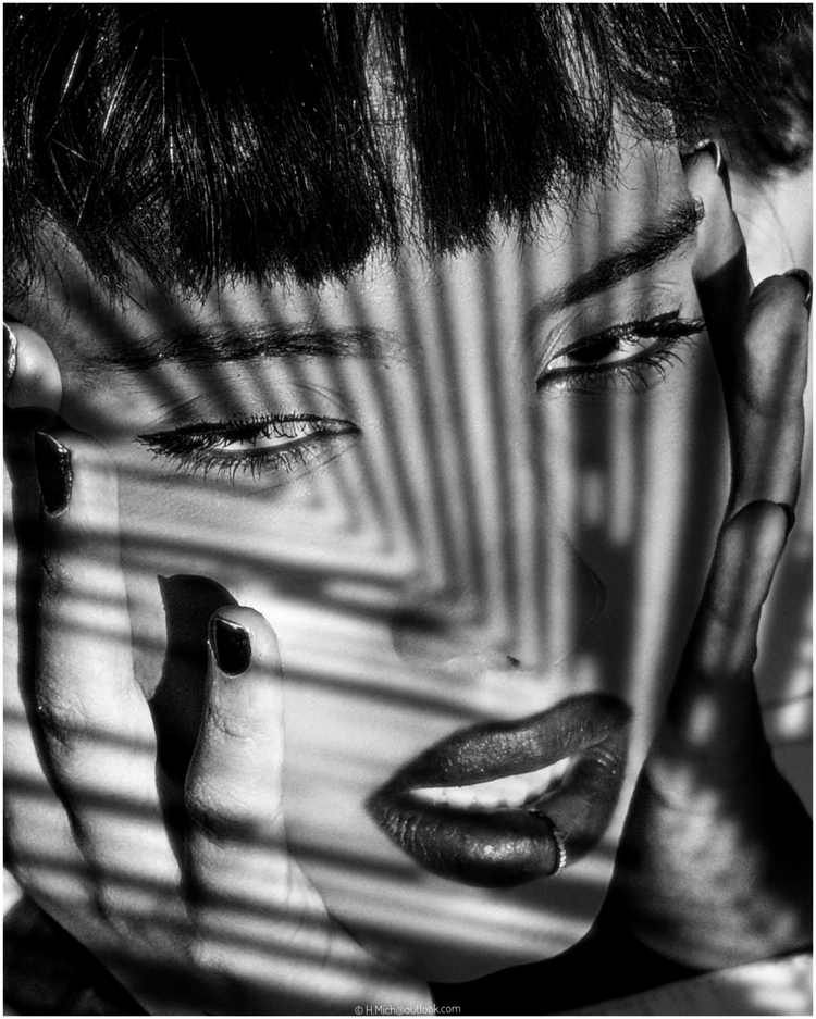 Diarium.Studio-One.Art.Br Black and white photo of a person with dark lipstick framed by hands, shadowed by striped light patterns, possibly from blinds. Eyes are partially closed, creating an intense and artistic effect. (c) H.Mich