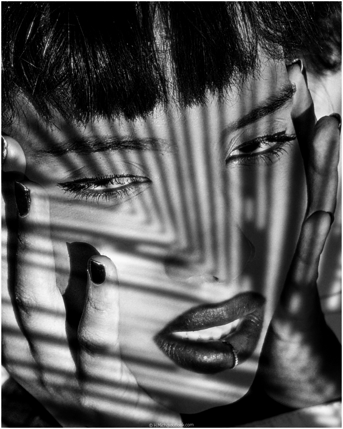 Diarium.Studio-One.Art.Br Black and white photo of a person with dark lipstick framed by hands, shadowed by striped light patterns, possibly from blinds. Eyes are partially closed, creating an intense and artistic effect. (c) H.Mich
